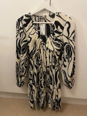 Plisserad klänning med svartvitt mönster - Snygg plisserad klänning från H&M med svartvitt blommigt mönster. Klänningen har v-ringning, långa puffärmar och en lös, flowy passform. 