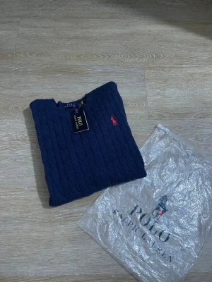Mörkblå kabelstickad tröja Polo Ralph Lauren - Mörkblå kabelstickad tröja från Polo Ralph Lauren med rund halsringning och röd broderad logga på bröstet. Tröjan har långa ärmar och ribbstickade muddar. Klassisk och stilren design, perfekt för lager-på-lager.