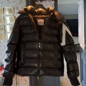 Svart dunjacka från Moncler - Säljer en svart glansig dunjacka från Moncler med vit detalj och logga på ena ärmen. Jackan har hög krage, dragkedja framtill och breda kanaler fyllda med dun. Perfekt för kalla vinterdagar och riktigt snygg streetstil.