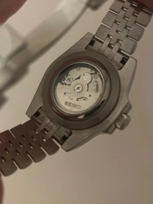 Seiko mod Bruce Wayne  - Snygg automatisk klocka från med robust länkarmband i silverfärgat stål. Urtavlan är svart med tydliga siffror och visare, samt datumfönster. Baksidan har ett transparent fönster som visar urverket. Perfekt för dig som gillar klassisk och sportig stil.