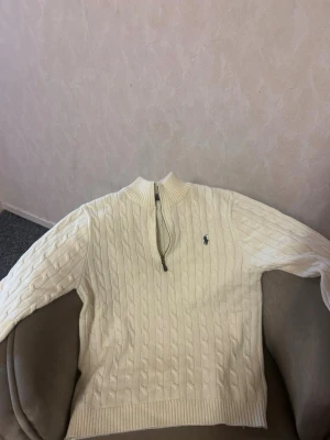 Vit kabelstickad tröja Polo Ralph Lauren - Vit kabelstickad tröja från Polo Ralph Lauren med halv dragkedja och broderad logga på bröstet. Tröjan har ribbade muddar och hög krage, perfekt för en stilren och klassisk look.