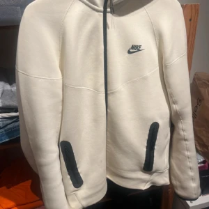 Ljusgul hoodie från Nike med svarta detaljer - Snygg ljusgul hoodie från Nike med svart logga på bröstet och svarta dragkedjor på fickorna. Tröjan har huva med snörning och är långärmad. 