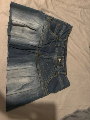Kort jeanskjol med slitningar - Säljer en cool kort jeanskjol i blå denim med slitna detaljer och markerade sömmar. Kjolen har flera fickor fram och bak samt en knappstängning framtill. Perfekt för dig som gillar en avslappnad och trendig stil.