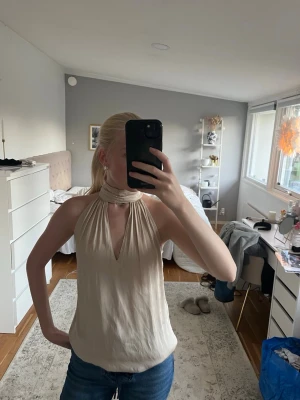 Champagnefärgad halterneck topp med knyt - Säljer en snygg halterneck topp med knyt runt halsen och djup v-ringning. 