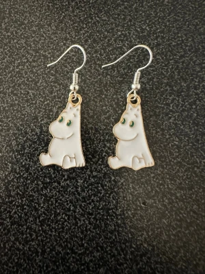 Mumin örhängen i vit emalj - Supergulliga örhängen med Mumin-motiv i vit emalj och gröna detaljer för ögonen. Hängande modell med silvriga krokar, perfekta för dig som älskar Mumin och vill ha något unikt till din stil. Krokarna är inköpta som nickelsäkra.