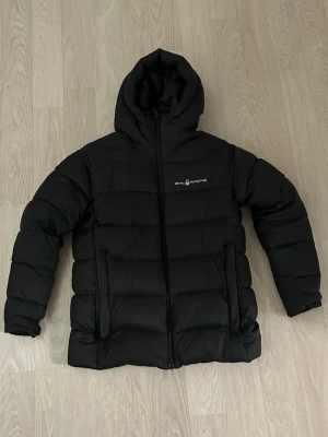 Svart pufferjacka från Sail Racing - Säljer en svart pufferjacka från Sail Racing med huva och dragkedja framtill. Jackan har en  diskret logga på bröstet och två sidofickor. Perfekt för kalla dagar och riktigt skön att bära. Den är i använt skick. Den är i storlek 170 för barn. Djur och rökfritt hem 