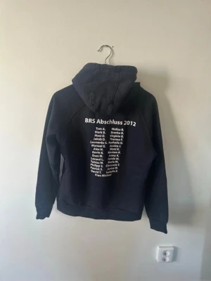Mörkblå Abschluss 2012 hoodie - Mörkblå hoodie med tryck 'Abschluss 2012' och stjärndesign framtill. Text och namn på ryggen samt tryck på ena ärmen. Klassisk känguruficka och justerbar huva med snören. Perfekt för dig som gillar unik och personlig stil.