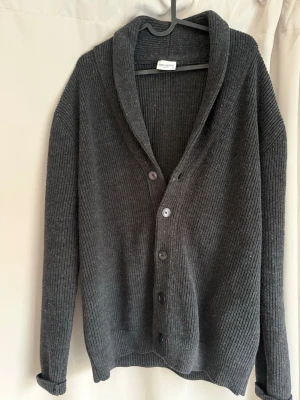 Mörkgrå ribbstickad kofta från john henric - Säljer en mörkgrå ribbstickad kofta från john henric med sjalkrage och knäppning framtill. Koftan har långa ärmar och en avslappnad passform, perfekt för lager på lager. Materialet är mjukt och ger en skön känsla.
