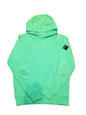 Grön hoodie från Stone Island - Fräsch grön hoodie från Stone Island med klassisk huva och ribbade muddar. Ikoniska Stone Island-märket sitter på vänster ärm. Tillverkad i mjukt bomullsmaterial som känns skönt mot huden. Perfekt för dig som vill ha en clean och stilren look.