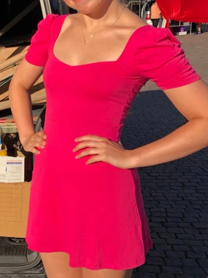 Rosa klänning - Snygg kort rosa klänning med små puffärmar och markerad midja. Klänningen har en lätt utställd kjol och en djup, fyrkantig ringning fram och djup ringning i ryggen. Materialet är stretchigt.