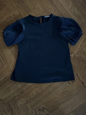 Snygg blå puffärmad topp, storlek 140 cm 🤩🩷 - Stilren svart topp med korta puffärmar i glansigt material. Rund halsringning och knapp i nacken. Perfekt för dig som vill ha en trendig och elegant look.