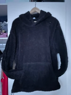 Svart fluffig hoodie - Säljer en svart hoodie i fluffigt teddy-material med huva och lång ärm. Hoodien har en avslappnad passform och en stor ficka framtill. Perfekt för dig som gillar mjuka och bekväma plagg med en trendig look.
