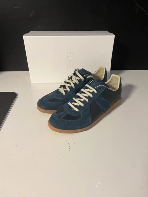 Blå sneakers från Maison Margiela - Snygga blå sneakers från Maison Margiela med ovandel i mocka och läder, vita skosnören och beige innersula. Skorna har en klassisk låg siluett och gummisula som ger en retrovibe. Perfekta för dig som gillar stilrena och tidlösa sneakers.