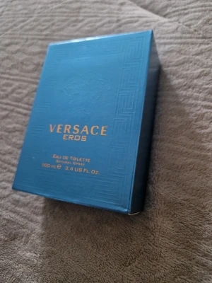 Versace Eros Eau de Toilette 100ml - Versace Eros Eau de Toilette i en snygg turkosblå glasflaska med guldiga detaljer och Medusa-logga. Flaskan rymmer 100 ml och kommer i en matchande blå kartong med guldtext. En ikonisk doft från Versace som sticker ut i badrumshyllan.