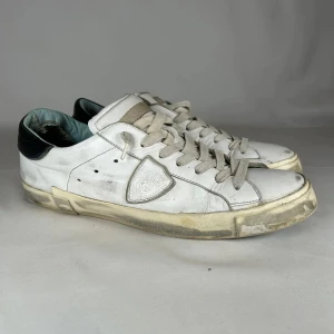 Vita sneakers med svart häl - Säljer ett par vita sneakers från Philippe Model med svart detalj på hälen och gräddfärgad sula. Skorna har snörning, rund tå och är tillverkade i skinn. Klassisk logotyp på sidan och låg profil som passar till många stilar.