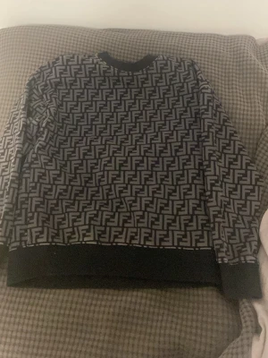 Svart mönstrad tröja från Fendi - Säljer en svart långärmad tröja från Fendi med det ikoniska FF-mönstret över hela plagget. Tröjan har rund halsringning och ribbade muddar vid ärmslut och nederkant. Perfekt för dig som gillar lyxiga streetwear vibes.