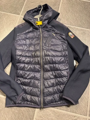 Mörkblå pufferjacka från Parajumpers - Säljer en mörkblå pufferjacka från Parajumpers med huva och snygga quiltade paneler framtill och baktill. Jackan har dragkedja, bröstficka med zip och ikonisk gul detalj vid halsen. Perfekt för kyliga dagar och riktigt skön passform. Nyskick storlek Small