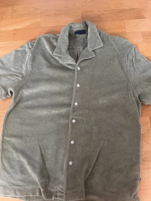 Grön frotté t-shirt från Lexington - Säljer en grön t-shirt från Lexington i mjuk frotté med korta ärmar och knappar framtill. Skön krage och relaxed fit, perfekt för chill dagar. Klassisk och enkel design som funkar till det mesta.