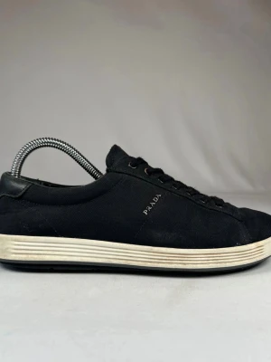 Svarta sneakers från Prada - Svarta sneakers från Prada med klassisk rund tå och platt vit sula. Skorna har snörning och en röd detalj på plösen. Tillverkade i textil med skinnpanel vid hälen. Diskret Prada-logga på sidan. Perfekt för dig som gillar stilrena och lyxiga sneakers.