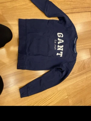 Mörkblå sweatshirt från GANT S - Säljer en mörkblå sweatshirt från GANT i storlek S. Tröjan har klassisk rund hals, långa ärmar och stor vit GANT-logga med 'Est. 1949' tryckt på bröstet. Tillverkad i mjuk bomull för skön vardagsstil.