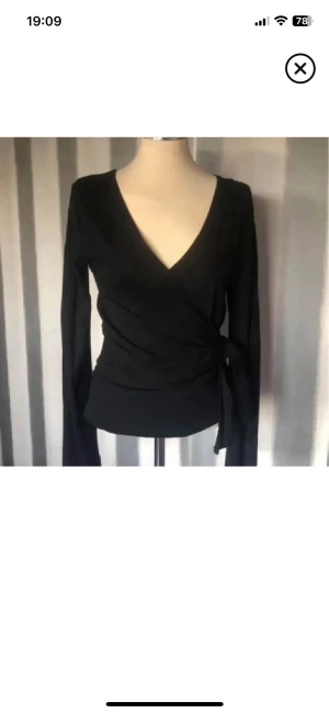 Svart omlottopp med knyt - Snygg svart omlottopp med långa ärmar och knyt i sidan. Djup v-ringning framtill ger en stilren look. Perfekt att matcha med jeans eller kjol för en trendig outfit.fin kvalitet 