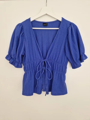 Blå knytblus från Gina Tricot XS - Supersöt blå blus från Gina Tricot i storlek XS. Blusen har puffiga korta ärmar, v-ringning och knytband framtill. Materialet är strukturerat och känns mjukt mot huden. Perfekt till jeans eller kjol för en trendig look. Resåren på ena armen har släppt något, men annars i fint skick.