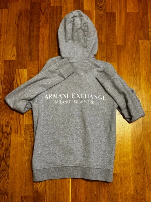 Armani Hoodie - Säljer en stilren grå hoodie från Armani Exchange med logga tryckt på bröstet och stor text på ryggen. Tröjan har huva med snörning och är långärmad. Perfekt för dig som gillar en clean och modern look.