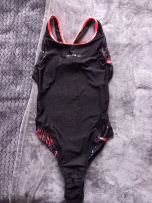 Svart baddräkt från Nabaiji - Säljer en svart baddräkt från Nabaiji med tunna korsade axelband i orange. Sidorna har coola grafiska detaljer i rött och vitt. Materialet är stretchigt och snabbtorkande, perfekt för simning. Snygg och sportig look med diskret mönster på framsidan.