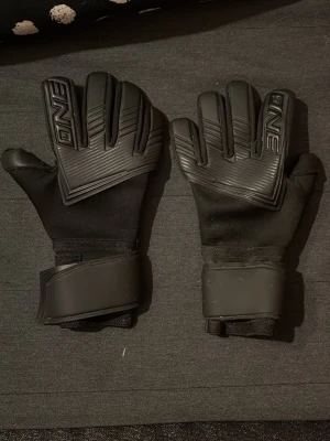 Svart ONE målvaktshandskar strl 9 - Säljer ett par svarta målvaktshandskar från ONE i storlek 9. Handskarna har en clean look med präglade detaljer på ovansidan och breda kardborreband vid handleden för extra stöd. Perfekta för dig som vill ha stilrena handskar på planen.