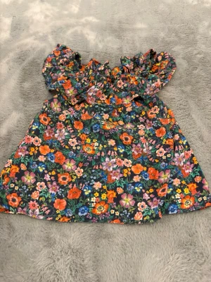 Blommig offshoulder topp från H&M - Supersöt offshoulder topp från H&M i storlek 140, täckt av färgglada blommor i orange, rosa, blått och lila på mörk botten. Toppen har volangdetaljer runt axlarna och är perfekt för sommarens alla äventyr.