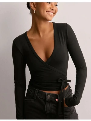 Svart omlottopp från Nelly - Säljer en svart omlottopp från Nelly i storlek XXS. Toppen har lång ärm, djup v-ringning och knyt i midjan för en snygg passform. Perfekt att styla med jeans eller kjol för en trendig look.