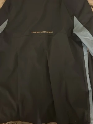 Under Armour svart vindjacka - Snygg svart vindjacka från Under Armour med ljusblå detaljer längs ärmarna. Jackan har dragkedja framtill, huva med snörning och logga på bröstet. Perfekt för dig som vill ha en sportig look och gillar funktionella plagg.