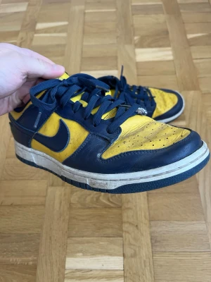 Nike Dunk Low blå och gula sneakers - Säljer ett par Nike Dunk Low sneakers i blått och gult skinn med klassisk vit och blå sula. Skorna har snörning och perforerade detaljer på tån för extra ventilation. Swoosh-logga på sidan och vadderad plös för skön passform. Perfekta för dig som gillar färgstarka sneakers.