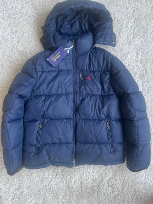 Mörkblå pufferjacka från Polo Ralph Lauren - Säljer en mörkblå pufferjacka från Polo Ralph Lauren med huva och dragkedja.     Skriv vid frågar.   Skickar alltid paketet inom 24 timmar