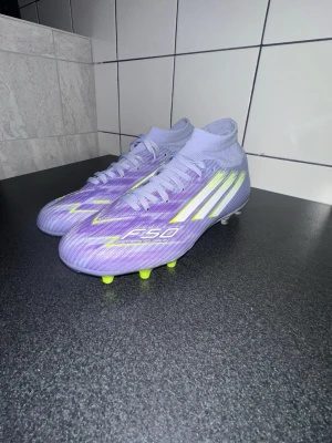 Adidas F50 Sparkfusion - Säljer ett par lila Adidas F50 fotbollsskor med vita och neongula detaljer. Skorna har snörning, högre strumpkrage och mönstrad sula med gula dobbar. Perfekta för dig som vill sticka ut på planen med snygg design och grym komfort.