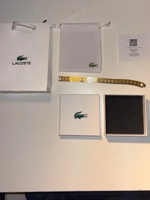 Guldigt armband från Lacoste - Snyggt guldigt armband från Lacoste med länkdesign och ingraverad krokodil på spännet. Armbandet har en stilren och modern look med detaljerade mönster på länkarna. Kommer med originalpåse, ask och certifikat.