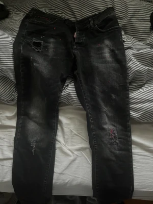 Svarta Dsquared2 jeans med slitningar - Säljer ett par svarta jeans från Dsquared2 med coola slitningar och färgstänk i rött och vitt. Jeansen har en rak passform och klassisk femficksdesign. Materialet är robust denim och de har en snygg, lite sliten look som sticker ut.