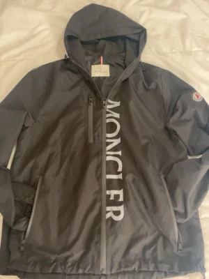 Svart vindjacka från Moncler - Snygg svart vindjacka från Moncler med stor logga längs dragkedjan och klassisk patch på ärmen. Jackan har huva, flera dragkedjeförsedda fickor och en clean look. 