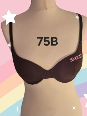 Brun Snoopy bh 75B - Mörkbrun bh med justerbara axelband och bygel, dekorerad med Snoopy-tryck i vitt och rosa på ena kupan. Klassisk tvåradig hyskknäppning bak och mjukt material som känns bekvämt mot huden. Perfekt för dig som gillar gulliga detaljer! Varan är oanvänd och kommer från ett konkurslager från 2000-talet. I många fall är prislappen kvar (se bild). Har fler annonser och bra bundlerabatt vid köp av flera.