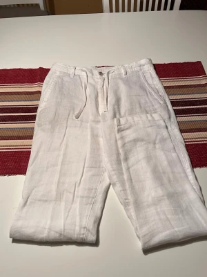 Vita linnebyxor med snöre från ZARA - Säljer ett par vita linnebyxor med avslappnad passform och snörning i midjan. Byxorna har sidofickor och en enkel knappstängning framtill. Perfekta för varma dagar och ger en chill vibe till din outfit. Från ZARA STORLEK 36. Knappt använt 