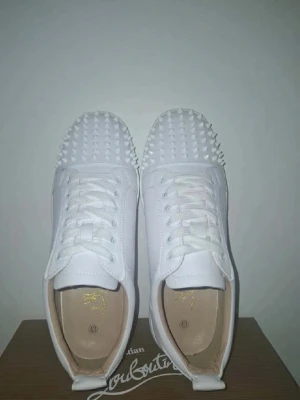 Vita Christian Louboutin sneakers med nitar - Säljer ett par vita sneakers från Christian Louboutin med ikoniska röda sulor och coola nitar över hela tån. Skorna har snörning och är tillverkade i skinn för en lyxig känsla. Perfekta för dig som vill sticka ut med exklusiv design och edgy detaljer.