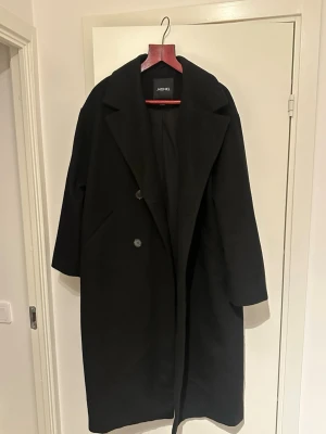 Svart lång kappa från Monki, stl M - Säljer en svart lång kappa från Monki i storlek M. Klassisk modell med bred krage, knappar framtill och fickor på sidorna. Kappan har en rak och loose passform, perfekt för höst och vinter. Materialet känns mjukt och värmande. Är lite använd, stylig och lite oversized. 