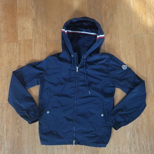 Moncler windbreaker - Marinblå windbreaker från moncler. Strlk 1 (S).          Självklart äkta,                                                         Defekter- saknar en metall bit som hänger frå ena snöret (bild 3), saknar även triangle zippen men annars så är allt som det ska vara. Pris kan diskuteras!