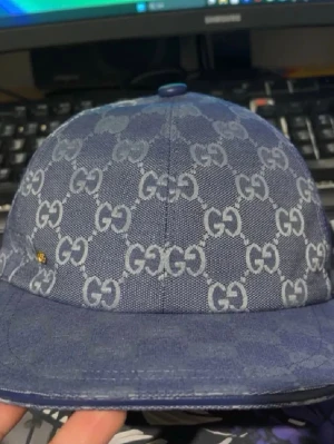 Blå Gucci keps med GG-mönster - Säljer en blå keps från Gucci med det klassiska GG-mönstret över hela kepsen. Kepsen har justerbart spänne bak i blått och en liten gulddetalj på sidan. Materialet är slitstarkt och har en snygg struktur. Perfekt för dig som vill ha en stilren accessoar.