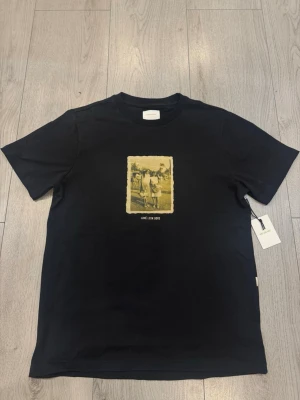 Svart Aimé Leon Dore t-shirt med print - Svart t-shirt från Aimé Leon Dore med ett retroinspirerat fotoprint i gult och vitt på bröstet. Klassisk passform och rund hals. Tillverkad i mjuk bomull som känns skön mot huden. Perfekt för dig som gillar streetwear och stilrena detaljer.