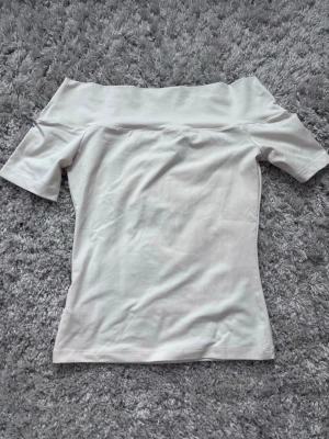 Vit offshoulder topp från H&M - Säljer en stilren vit offshoulder topp från H&M i mjukt bomullsmaterial. Toppen har korta ärmar och en bred kant upptill som ger en snygg axelbandslös look. Perfekt att matcha med jeans eller kjol för en clean och fräsch outfit.