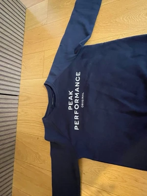 Mörkblå Peak Performance sweatshirt - Mörkblå sweatshirt från Peak Performance med vit logga tryckt på bröstet. Klassisk rund halsringning med ribbad kant och långa ärmar. Tillverkad i mjuk bomullsblandning, perfekt för chill dagar eller till skolan.