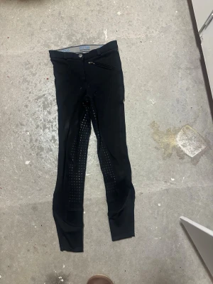 Svarta ridbyxor från CRW, stl 150 - Säljer ett par svarta ridbyxor från CRW Classic Riding Wear i storlek 150. Byxorna har helskoning med grepp i sätet och insidan av benen, bälteshällor och en dragkedjeförsedd ficka fram. Perfekta för dig som gillar ridning och vill ha bra grepp i sadeln.