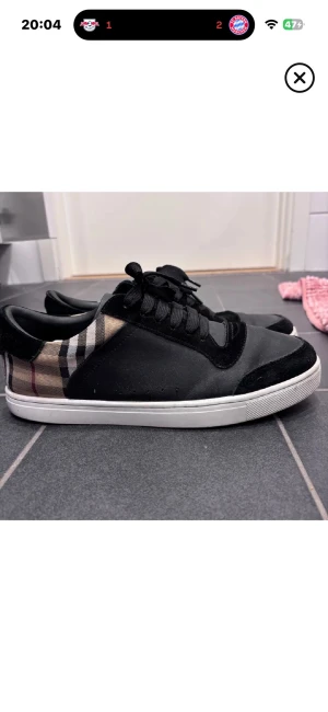Burberry svarta sneakers med rutigt mönster - Snygga svarta sneakers från Burberry med klassiskt beige, svart och rött rutigt mönster på hälen. Skorna har vita sulor, svarta snören och detaljer i mocka och skinn. Perfekta för dig som vill ha en stilren men ändå statement look.