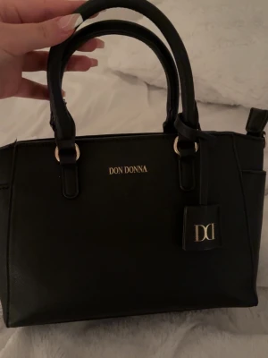 Svart handväska från Don Donna - Snygg svart handväska från Don Donna med guldfärgade detaljer och dubbla handtag. Klassisk och stilren design med logga framtill och en liten tag med DD-logga. Perfekt storlek för det viktigaste och enkel att matcha till olika outfits.